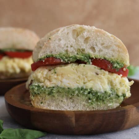 Cut open spinach pesto egg sandwich on a wood plate, showing the layers of pesto, eggs, cheese and tomatoes.