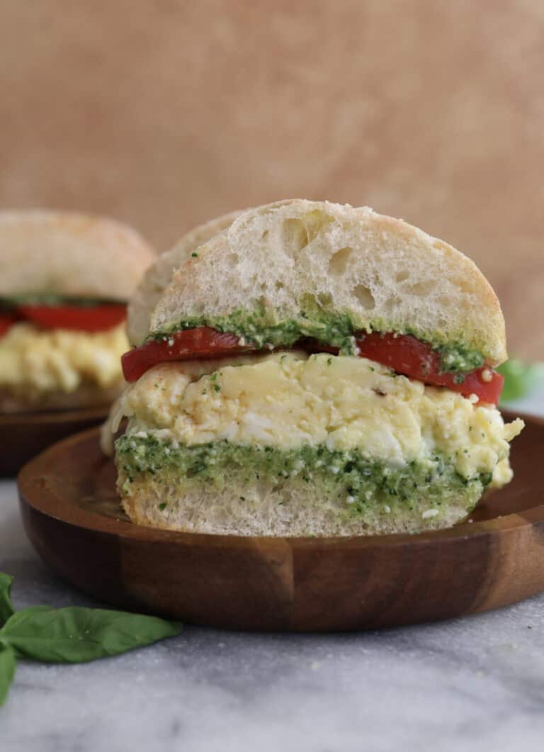 Cut open spinach pesto egg sandwich on a wood plate, showing the layers of pesto, eggs, cheese and tomatoes.