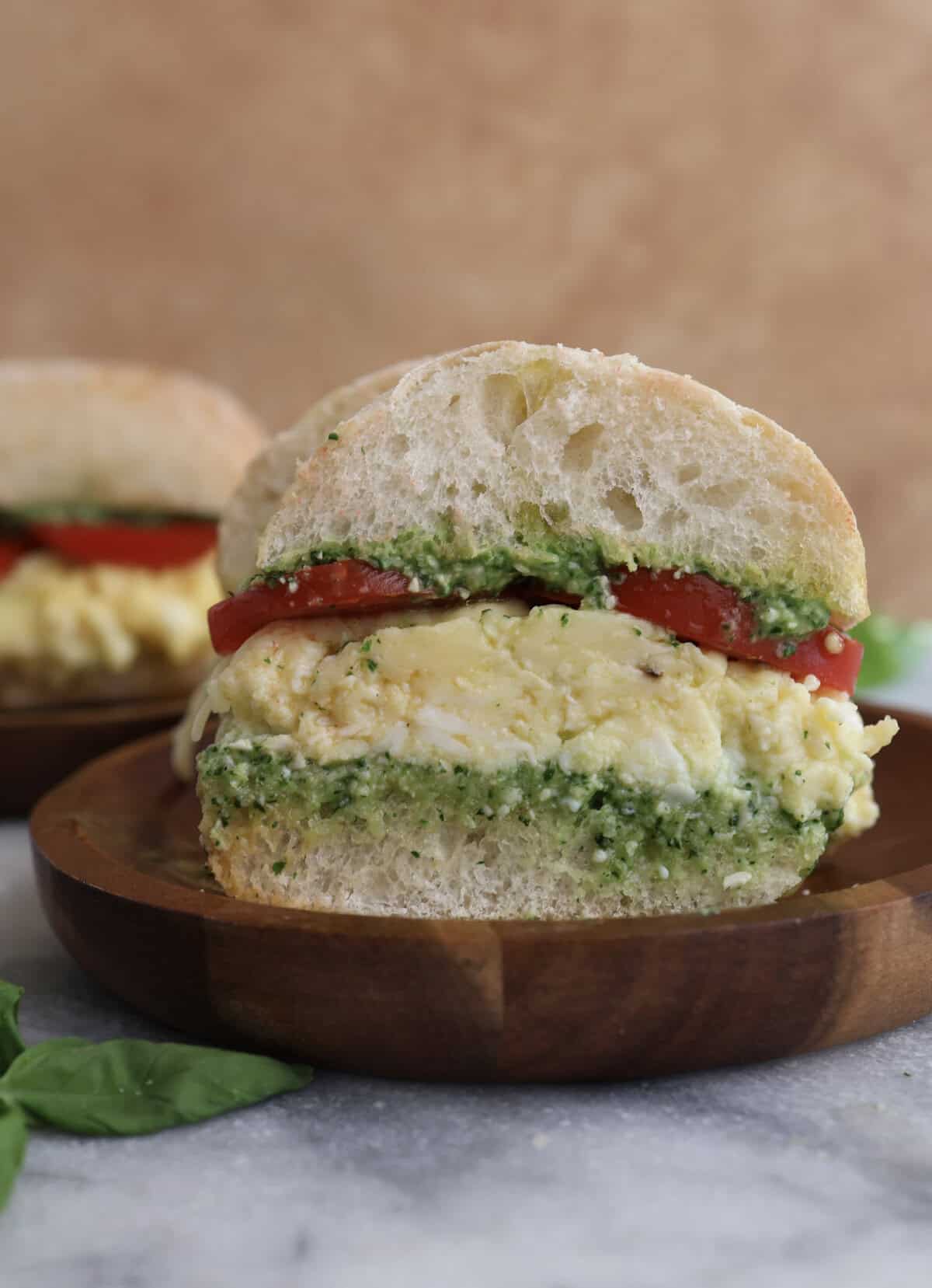 Cut open spinach pesto egg sandwich on a wood plate, showing the layers of pesto, eggs, cheese and tomatoes.