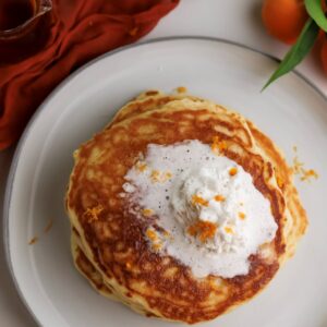 A stack of ricotta orange pancakes on plate with whipped cream.
