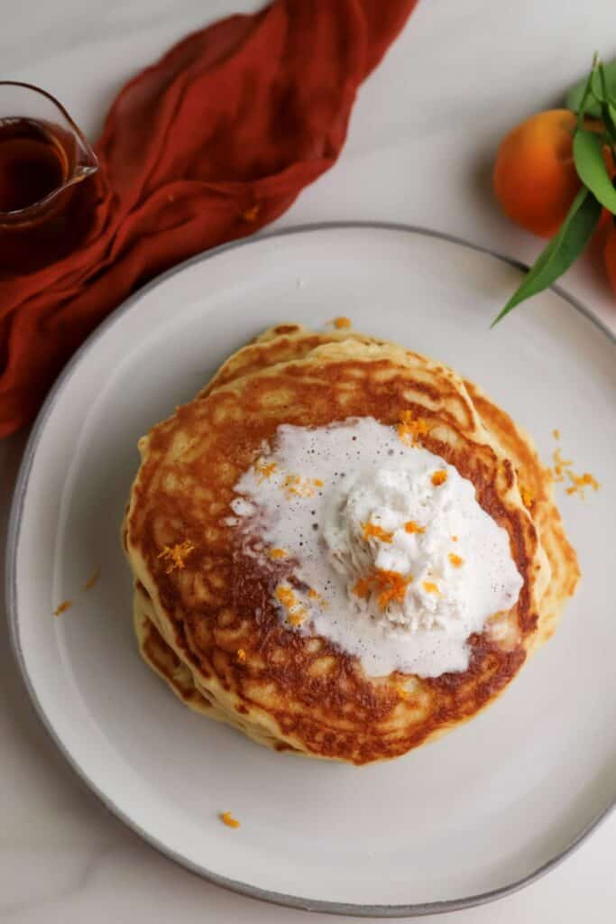 A stack of ricotta orange pancakes on plate with whipped cream.