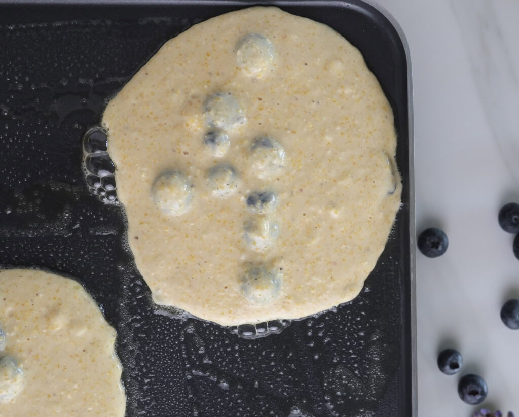 Cornmeal blueberry pancake batter on a griddle cooking.