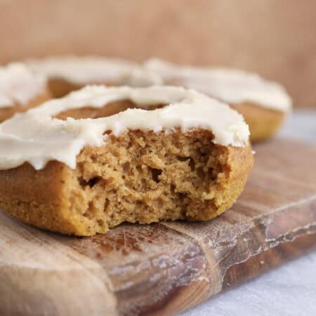 Baked pumpkin donut with maple icing sitting on a wood cutting board with a bite taken out, showing the soft texture of the donut.