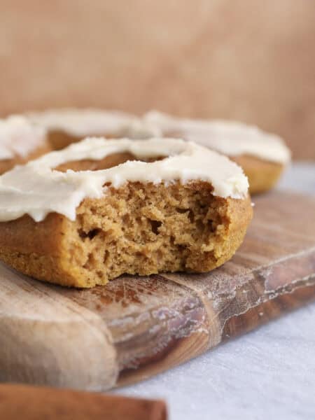 Baked pumpkin donut with maple icing sitting on a wood cutting board with a bite taken out, showing the soft texture of the donut.