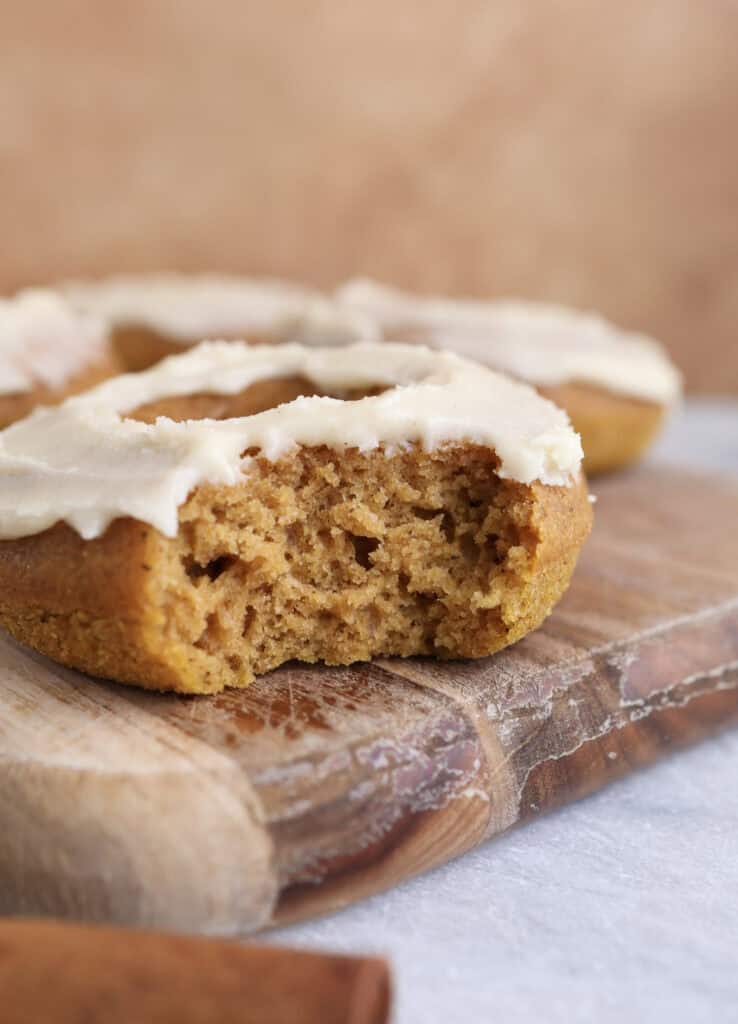 Baked pumpkin donut with maple icing sitting on a wood cutting board with a bite taken out, showing the soft texture of the donut.