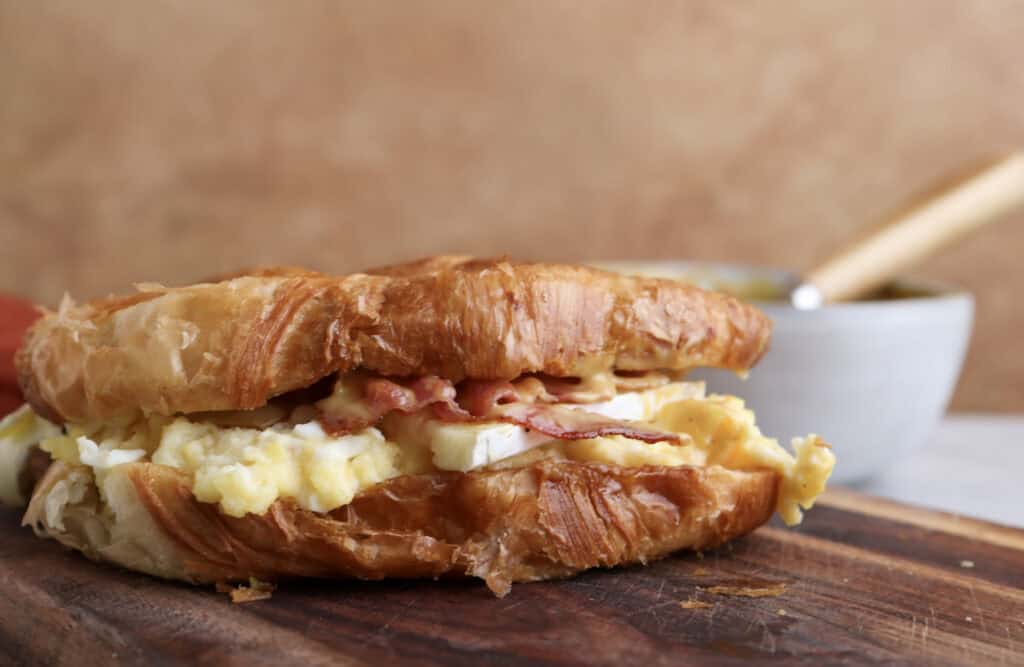 Breakfast croissant sandwich with bacon, egg, brie and spicy honey mustard on cutting board.