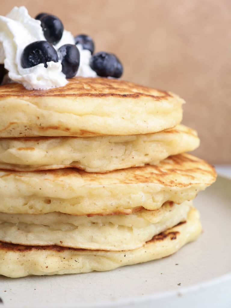 Up close shot of the side of the brown butte pancakes stacked on a plate topped with whipped cream and blueberries.