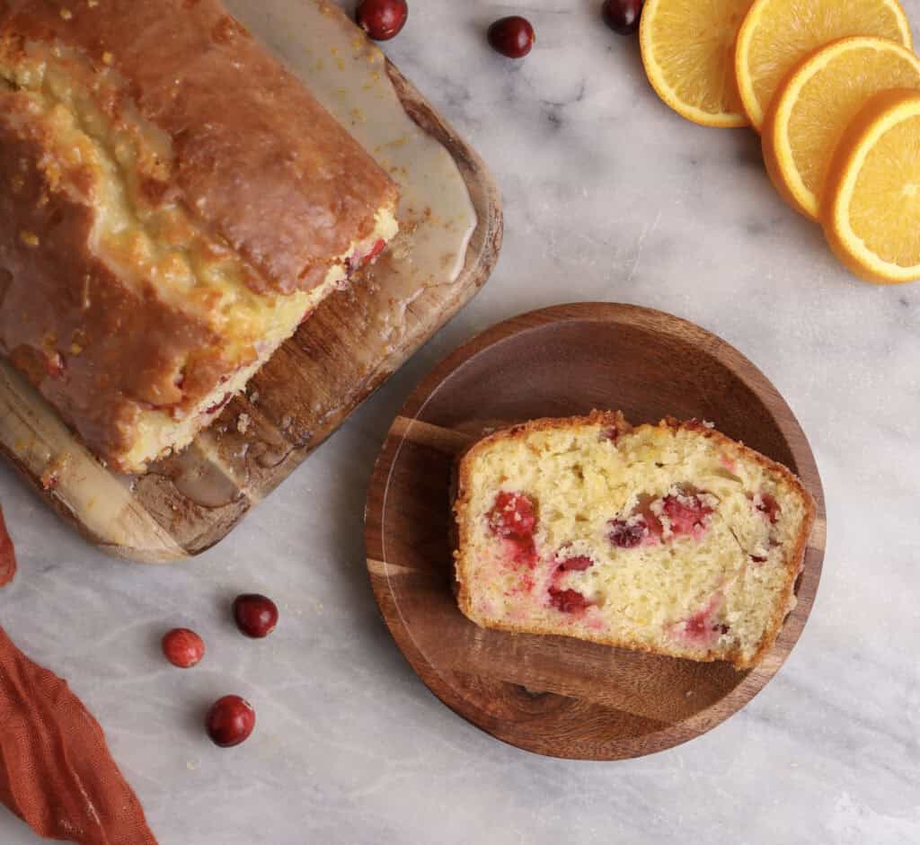 A slice of cranberry orange bread on a wood plate next to the loaf.