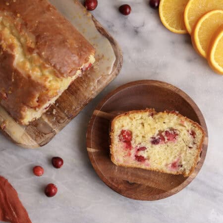 A slice of cranberry orange bread on a wood plate next to the loaf.