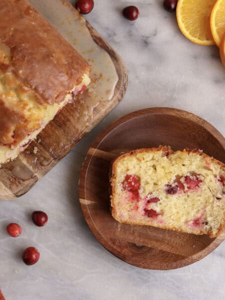A slice of cranberry orange bread on a wood plate next to the loaf.